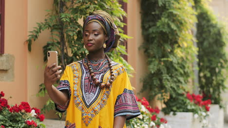 Portrait-Shot-Of-The-Young-Beautiful-Woman-In-The-Traditional-Clothes-Standing-In-The-Nice-Courtyard-With-Flowers-And-Having-Videochat-On-The-Smartphone