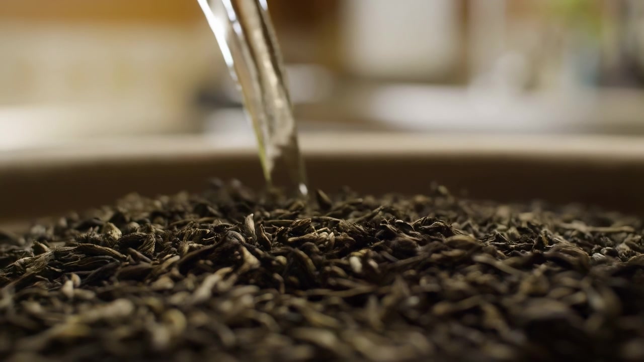 Clear water flows downward onto a bed of coffee beans, creating subtle movement across textured surfaces while warm light highlights the dark grains and close composition.