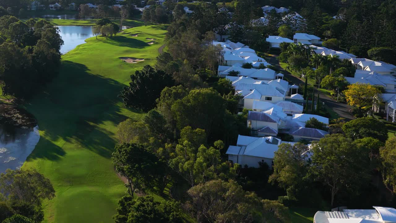 Drone footage captures a lush golf course beside a residential area under clear skies, showcasing vibrant greenery and serene surroundings