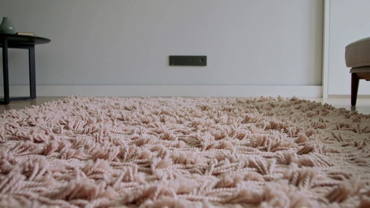 Kameraet trekkes tilbake og avslører et mykt, rosa rya-teppe i en moderne lounge, med et sidebord.