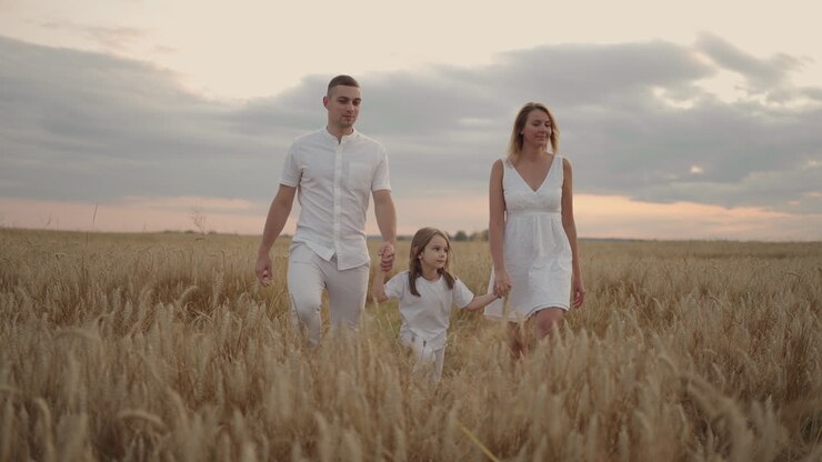 giovane coppia di genitori con bambine che si tengono per mano e corrono attraverso un campo di grano al tramonto. famiglia felice che corre tra un prato d'orzo e si gode la natura insieme. rallentamento
