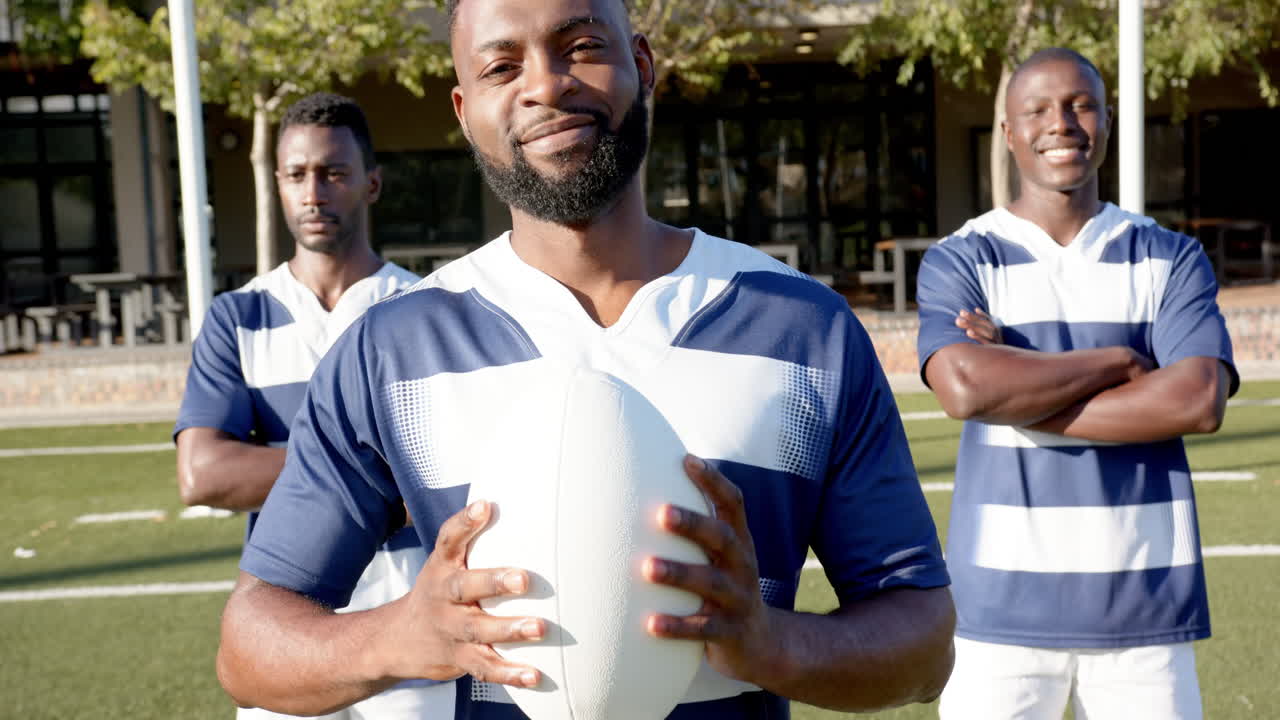 Homens afro-americanos segurando uma bola de rugby, com uniformes esportivos, em pé no campo, sorrindo.