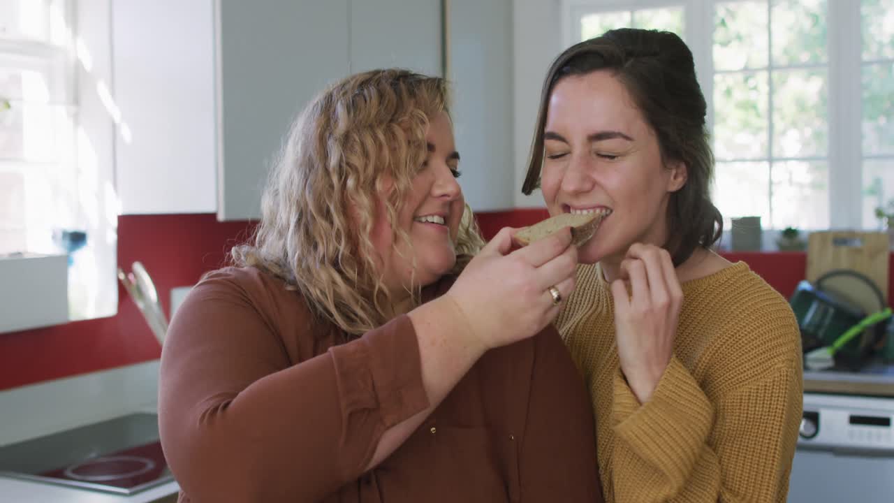 Happy caucasian lesbian couple eating bread and laughing in kitchen