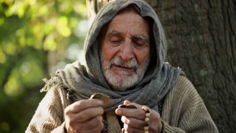 an elderly man sits calmly against a tree, holding a string of beads in his hands. sunlight filters through the leaves, creating a serene atmosphere. he appears thoughtful and content.