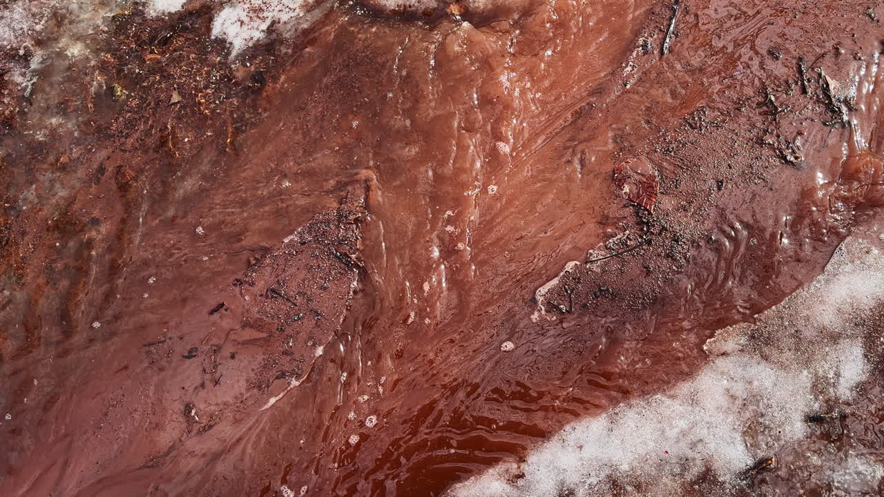 Chiara acqua di disgelo si insinua attraverso il terreno rosso che si scongela e il ghiaccio persistente, creando un primo piano riccamente strutturato che cattura il passaggio dall'inverno alla primavera.