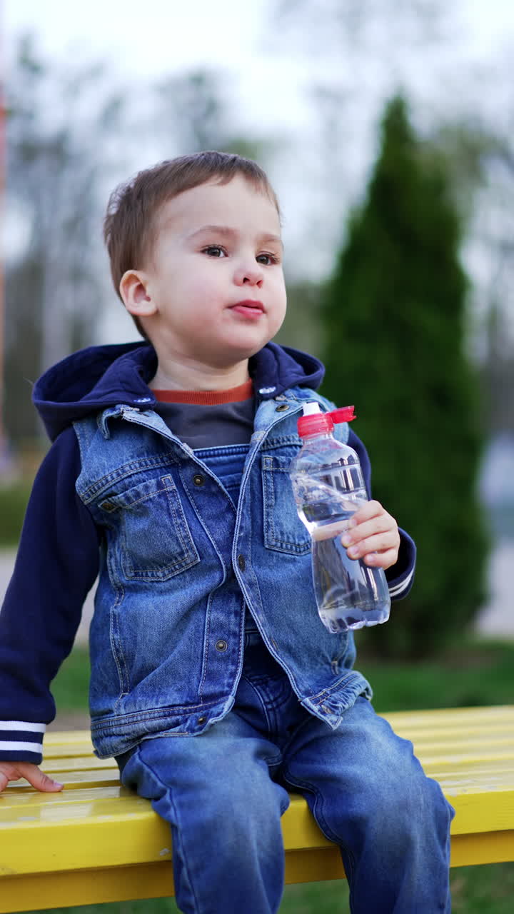 Kid wearing jeans clothes sits on the bench holding a bottle. Kid becomes angry as his mom cleans his nose with a napkin. Vertical video.