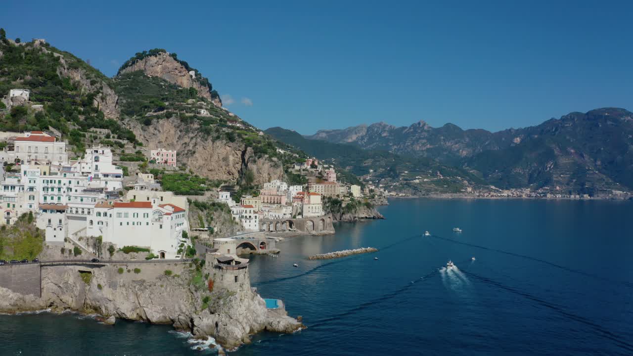 Ampia vista aerea con drone di una tortuosa strada di montagna lungo la Costiera Amalfitana attraverso le città di Amalfi e Atrani con barche che navigano lungo la costa del Mar Mediterraneo in una soleggiata giornata estiva