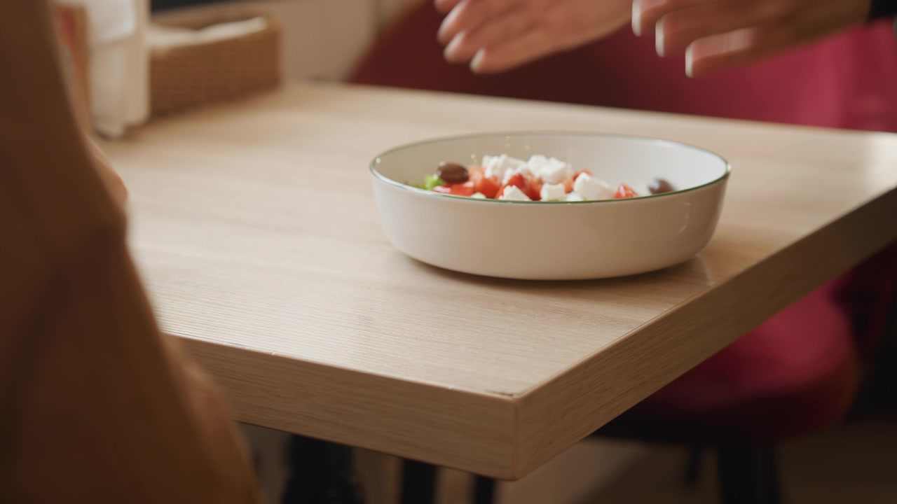 Close-up de mesa de madeira enquanto outra pessoa coloca e pousa uma tigela de salada. A cena apresenta iluminação suave, cadeiras vermelhas ao fundo e foca na interação entre duas pessoas com comida.