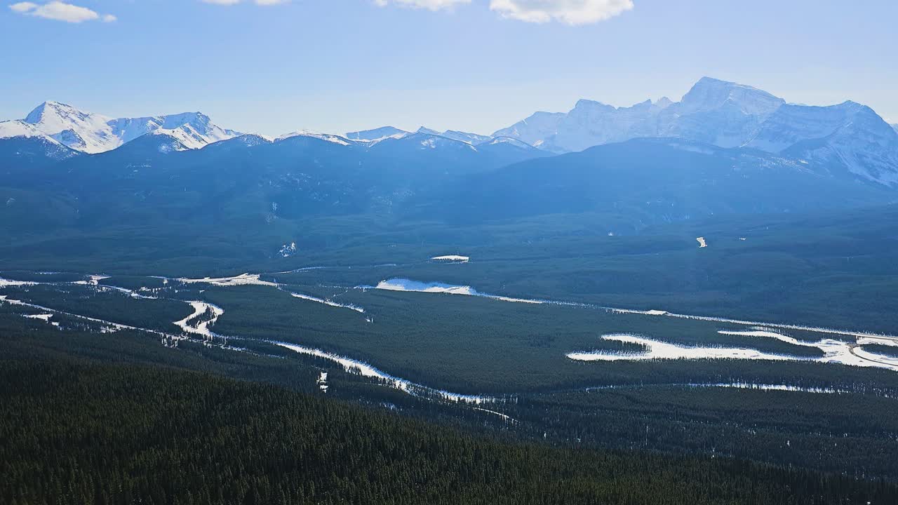 Paesaggio panoramico a Banff, Alberta, con aspre cime montuose, fitta foresta di conifere e un corso d'acqua serpeggiante sottostante, che crea una vista mozzafiato delle Montagne Rocciose canadesi alla luce del giorno