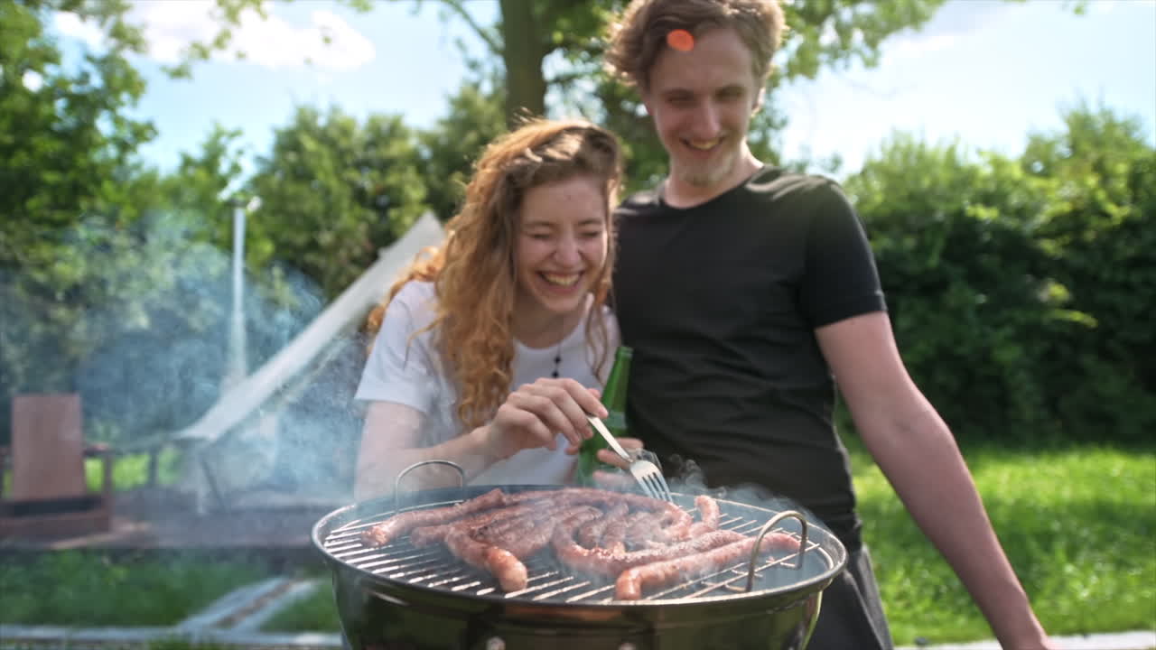 Giovane coppia che frigge carne alla griglia e beve birra. Tanto verde intorno. Glamping. Slow motion