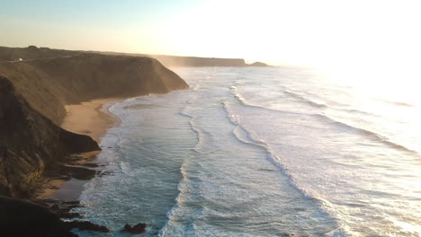 aerial drone shot of waves hitting a beach with cliffs during a sunset in algarve in southern portugal