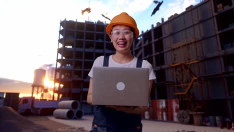 trabajadora asiática con gafas de protección y casco de seguridad usando una computadora portátil y sonriendo mientras está de pie en el sitio de construcción