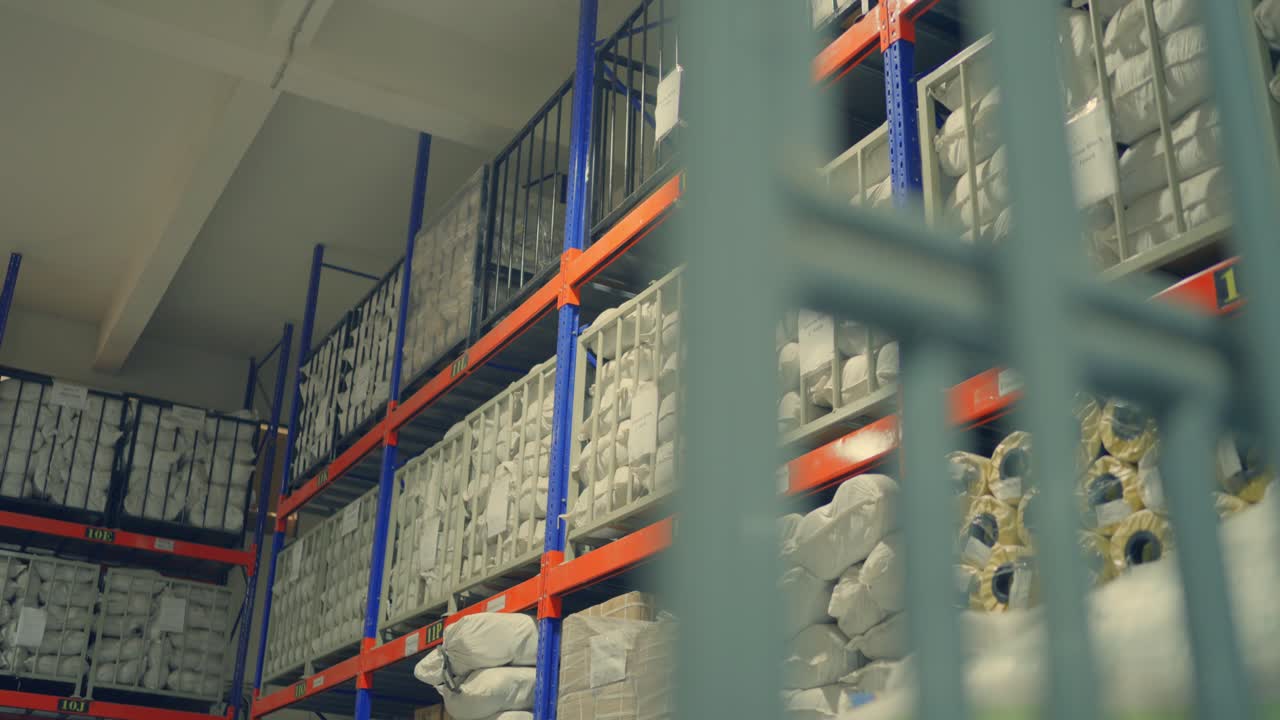 Movimento verso l'alto della telecamera all'interno di una fabbrica di tende Sialkot, che rivela scaffalature arancioni e blu dal pavimento al soffitto piene di inventario in magazzino