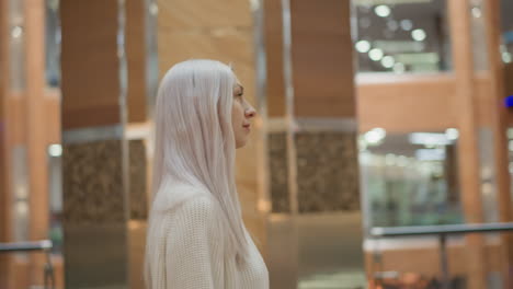 side view of elegant modern woman walking through urban mall corridor looking around in admiration under bright decorative lights against glass railings and polished floor exuding chic grace