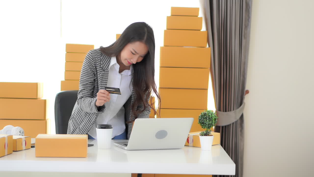 A woman entrepreneur, surrounded by shipping boxes in the background, hovers over her laptop, credit card in hand completing a transaction
