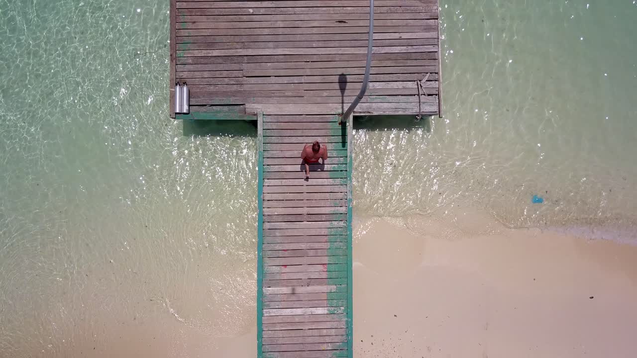 Man runs into frame and across pier then dives into bright blue waters on tropical island