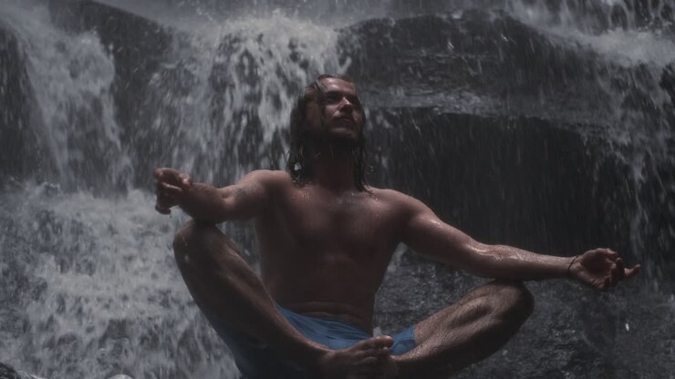 Young man meditating under waterfall.