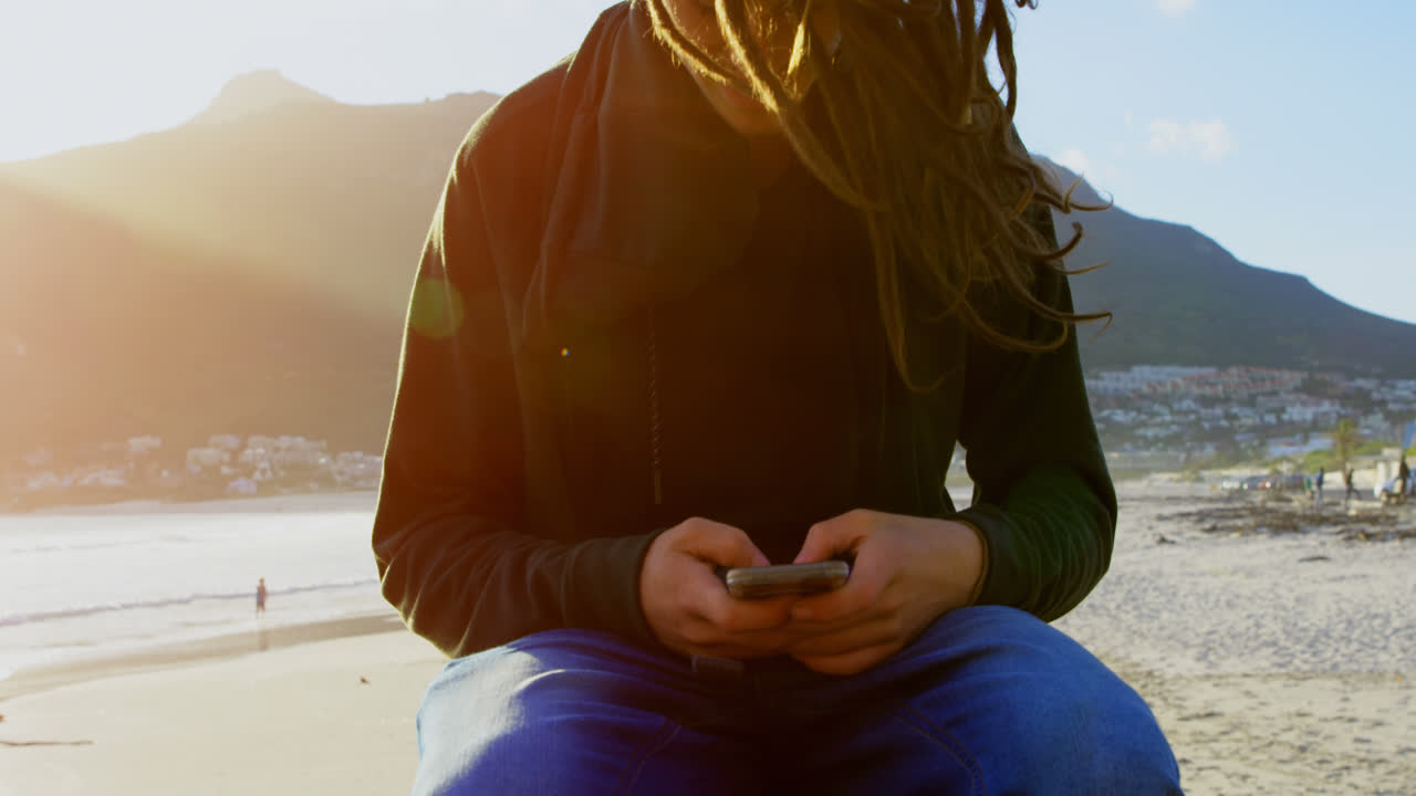 Vista frontale di un giovane uomo caucasico che scrive sul cellulare mentre è seduto su uno skateboard in spiaggia 4k