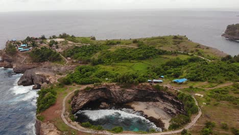 Filmische-Luftaufnahme-Der-Kreisförmigen-Felsformation-Am-Zerbrochenen-Strand-Mit-Meereswellen,-Die-An-Warmen,-Bewölkten-Tagen-Mit-Blauem-Wasser-In-Die-Mitte-Am-Rande-Der-Indonesischen-Insel-Stürzen