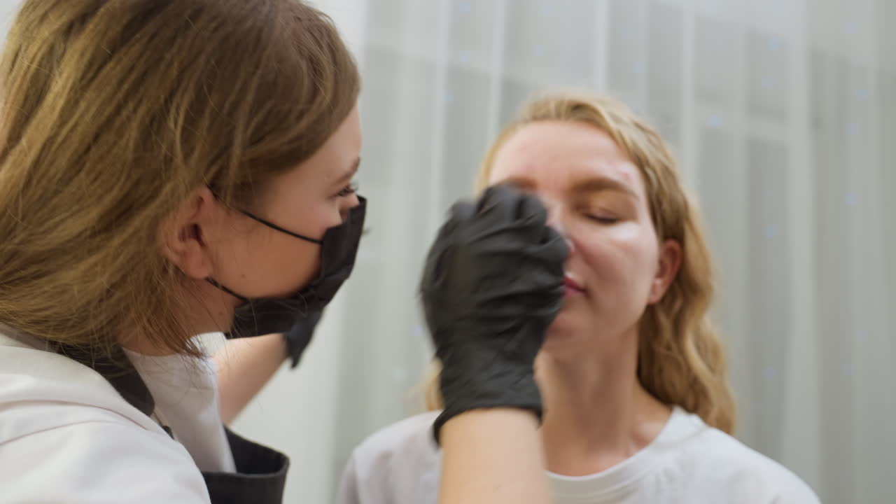 Beautician in gloves and face mask gently picks client eyebrow with tweezers during detailed grooming session. Client keeps eyes closed as she relaxes under soft lighting near white curtain backdrop
