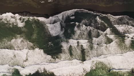 Sideways-aerial-top-down,-huge-waves-crashing-on-brown-sand-beach-in-Iceland