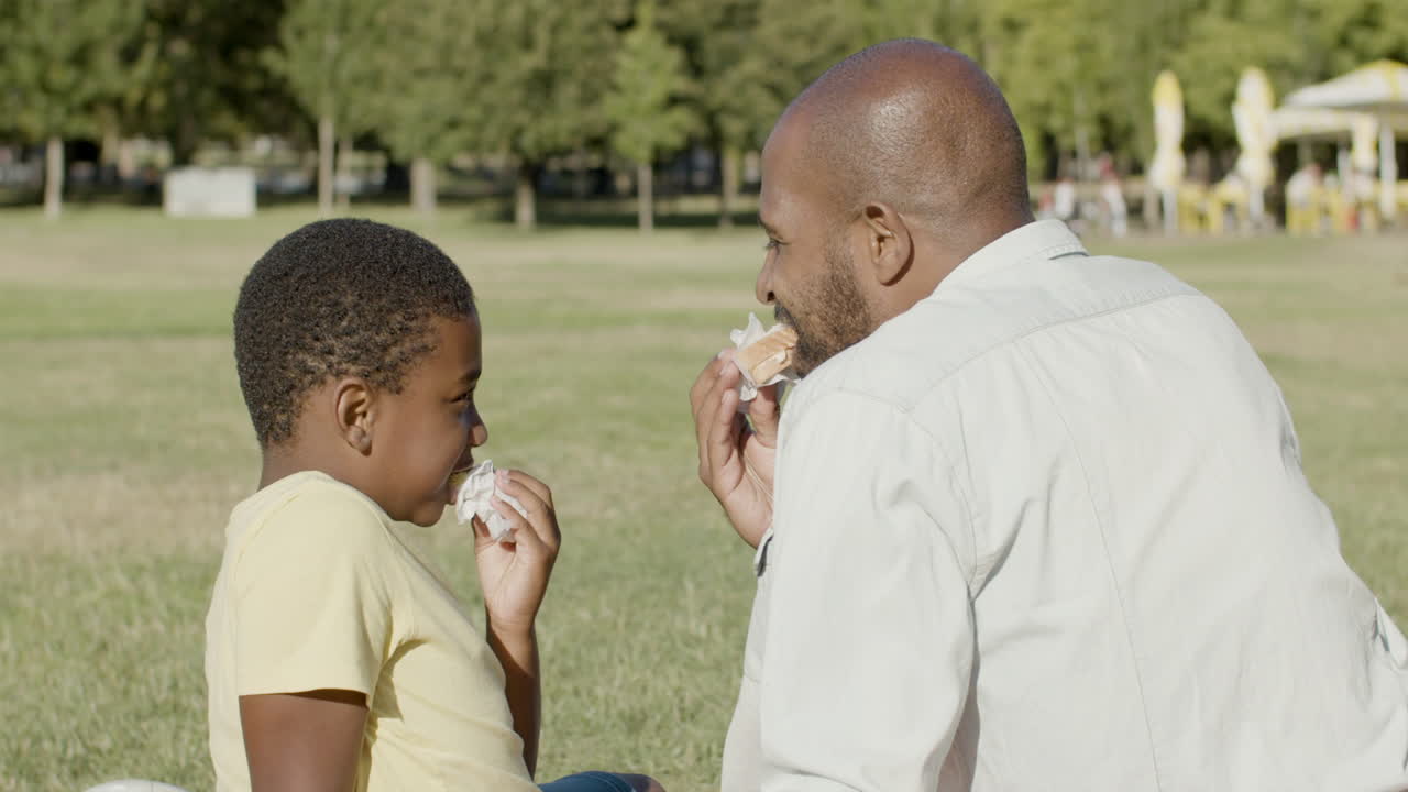 Padre e figlio che fanno un picnic al parco, mangiando panini.