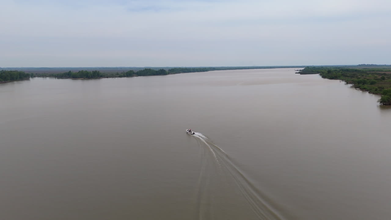 Motorboat speeds away from view across the broad muddy channels of the Paraná Delta, its twin white trails fanning toward distant tree-covered islets beneath a soft gray, cloud-filled sky.