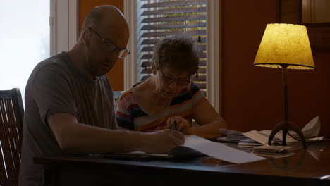a middle-aged man and a senior woman sit together at a dining table in warmly lit home, sorting through paperwork. woman dictates information, while man jots down notes in a notepad, with a back pen.
