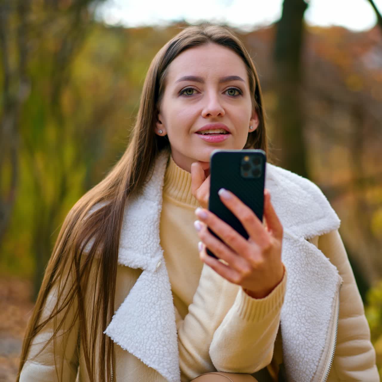 Donna caucasica con lunghi capelli scuri che trascorre del tempo nel parco autunnale. Donna con abiti leggeri e caldi che parla al telefono seduta al tavolo di legno