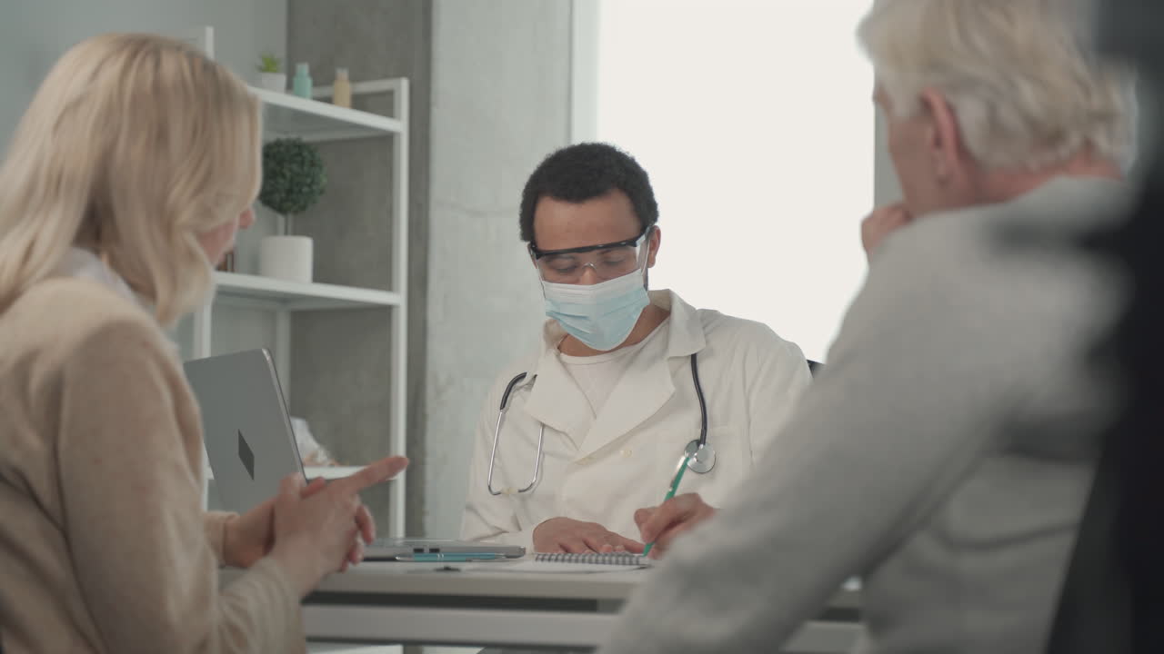 안면 마스크를 쓴 젊은 흑인 의사가 ederly 커플과 이야기하고 있습니다. 2