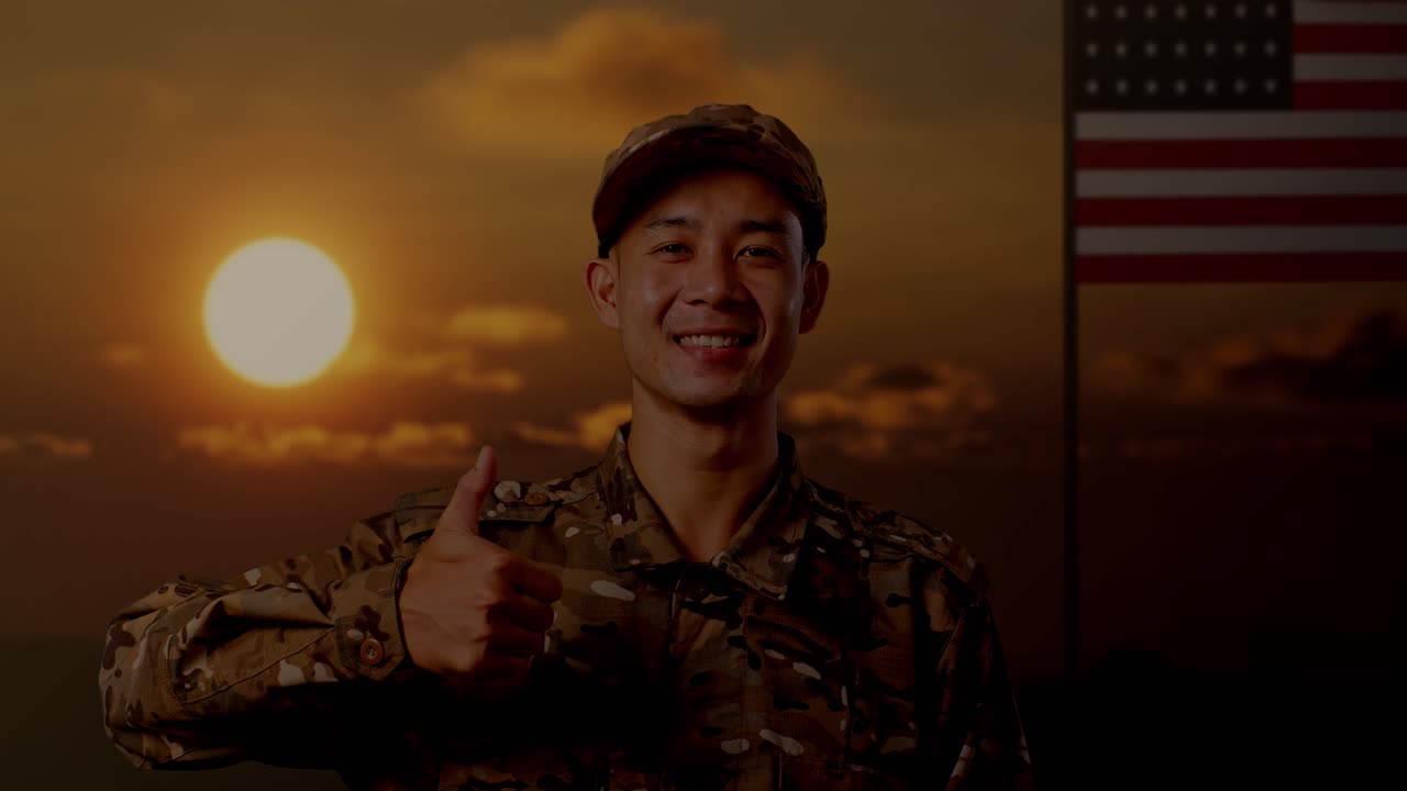 미소 짓고 엄지손가락을 올리는 아시안 남성 군인의 클로즈업 미국 발을 들고 서 있는 모습, 해가 지는 시간