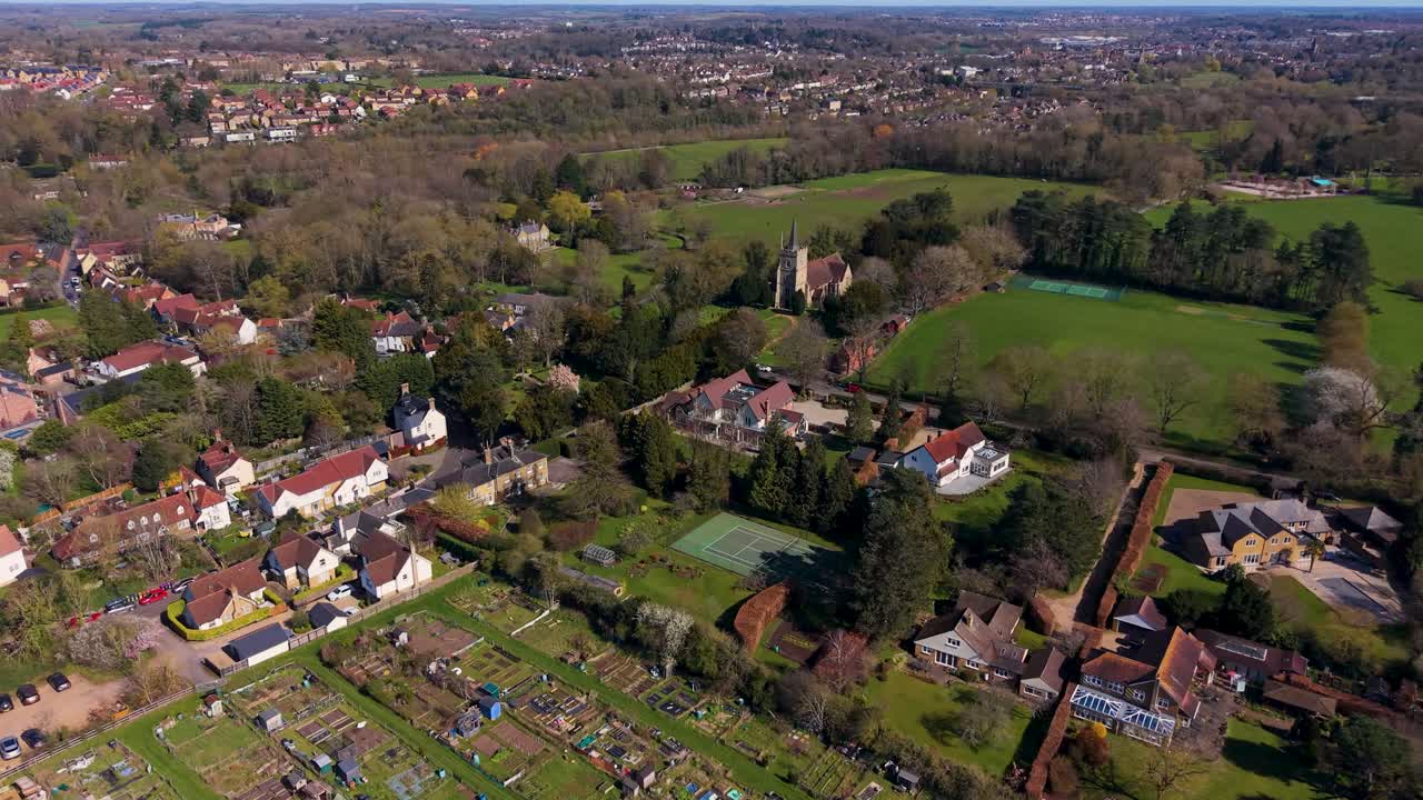 Luftfoto-kredsløb over landsbyen Hertingfordbury i Hertfordshire, England, der afslører forstadshuse, kirketårn, kolonihaver, træer og det omkringliggende landskab på en klar dag.