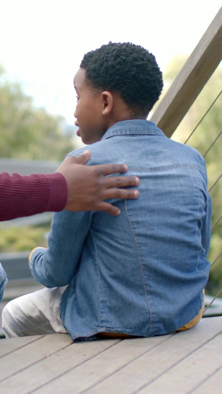 Vertical video: Maroon adult reaching left, placing hand on kid back on deck, comforting in denim