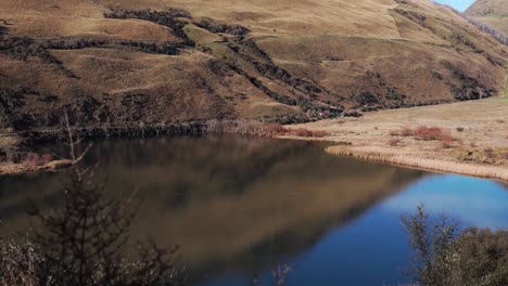 Perfectly-still-water-at-lake-surrounded-by-orange-rolling-hills-and-mountains-in-New-Zealand-countryside-causing-crisp-reflections