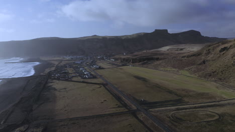 Pueblo-Costero-De-Vik-En-La-Playa-De-Arena-Negra-Con-Paisaje-Montañoso-Rocoso-En-El-Fondo-En-El-Sur-De-Islandia