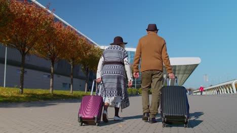 senior pensioner tourists grandmother grandfather walking to airport hall with luggage on wheels