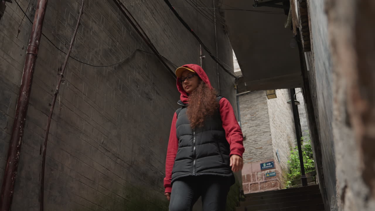 Mujer asiática descendiendo por un callejón estrecho de hormigón, con sudadera roja y chaleco negro, moviéndose bajo un saliente bajo. Paredes sombreadas con suciedad y tuberías, luz cinematográfica y melancólica, expresión introspectiva y solitaria.