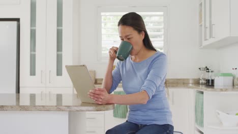 Feliz-Mujer-Asiática-Sentada-En-La-Mesa,-Tomando-Café-Y-Usando-Una-Tableta-En-La-Cocina