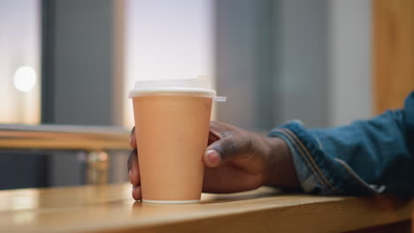 closeup of male hand gently holding takeaway coffee cup on wooden table during quiet moment indoors, soft daylight through window creates peaceful atmosphere, denim sleeve and warm light add cozy vibe