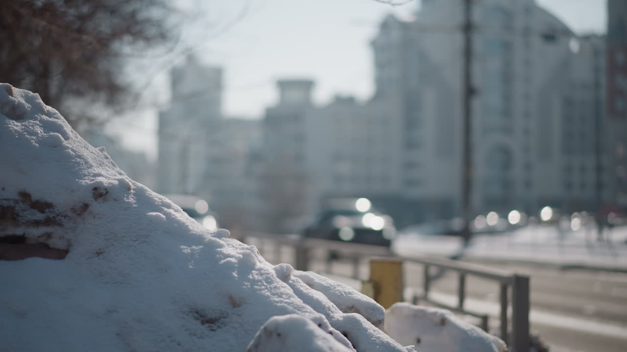 Close up snow mound by road under bright winter sun, blurred buses glide through city lane, headlights sparkle, tall buildings soften into bokeh as cold traffic flows past during midday commute