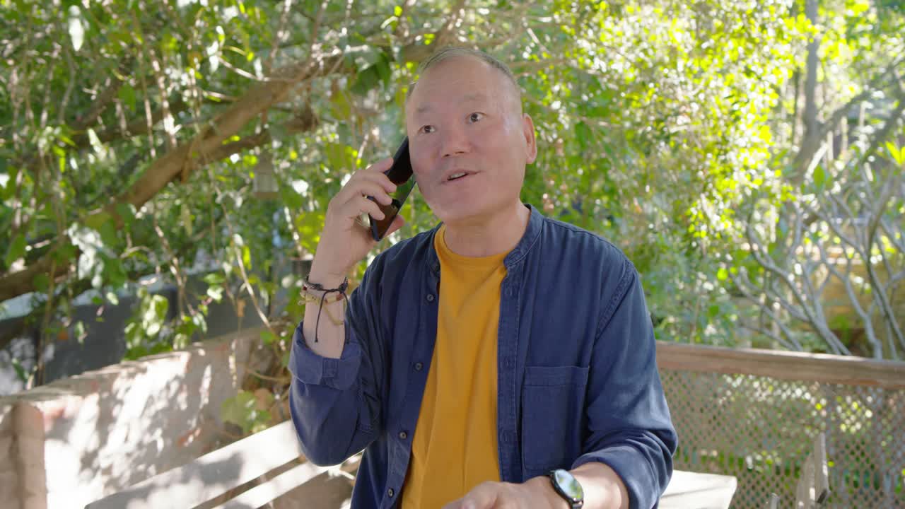 日当たりの良いテラスに立ち、スマートフォンを持ち、電話に応答しながらジェスチャーをする高齢のアジア人男性