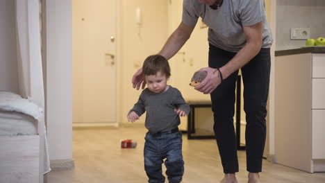 Father-Helping-Her-Baby-To-Take-His-First-Steps-From-The-Corridor-To-The-Living-Room-At-Home