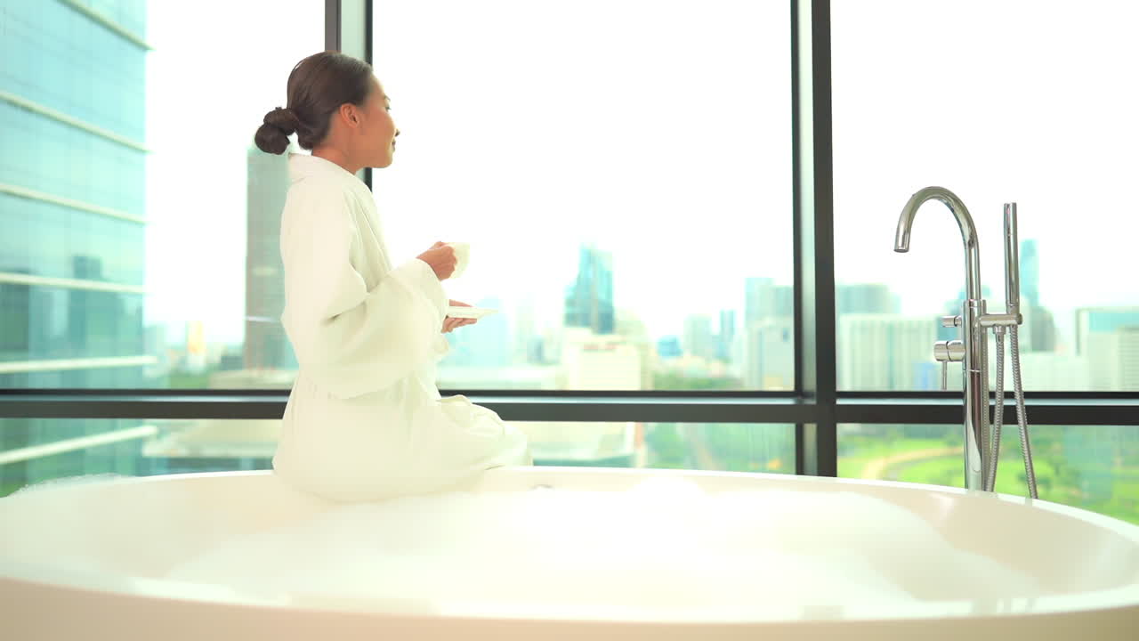 거품 목욕을 준비하는 동안 테리 가운을 입은 젊은 여성이 튜브 가장자리에 앉아 차 한 잔을 홀짝입니다.