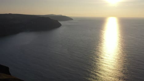 Drone-shot-tilting-up-while-looking-at-the-refection-of-sun-rays-on-the-ocean-from-sunset-on-the-North-Devon-coast,-looking-at-Lynton-and-Lynmouth-in-the-UK