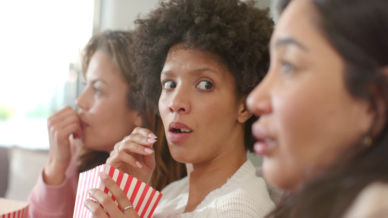 Women friends watching offscreen while eating popcorn tubs on sofa and central woman showing shock