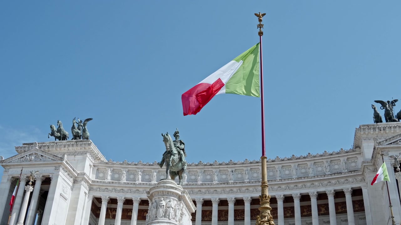 La bandiera tricolore italiana sventola sopra l'Altare della Patria e la statua equestre di Vittorio Emanuele II contro un cielo azzurro limpido