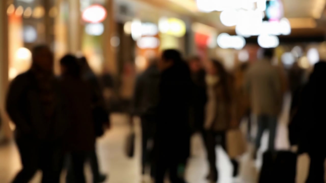 Folla sfocata in un centro commerciale con luci bokeh calde, sagome di pedoni e acquirenti in movimento.