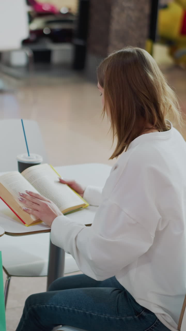 vista trasera de una mujer sentada en una mesa en un centro comercial leyendo un libro, rodeada de bolsas de compras, fondo borroso suave con personas que pasan por un centro comercial vibrante con luces