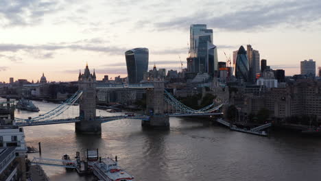 Cars-and-red-double-decker-driving-through-historic-Tower-Bridge-across-River-Thames-in-sunset-time.-Group-of-skyscrapers-in-City-business-centre.-London,-UK