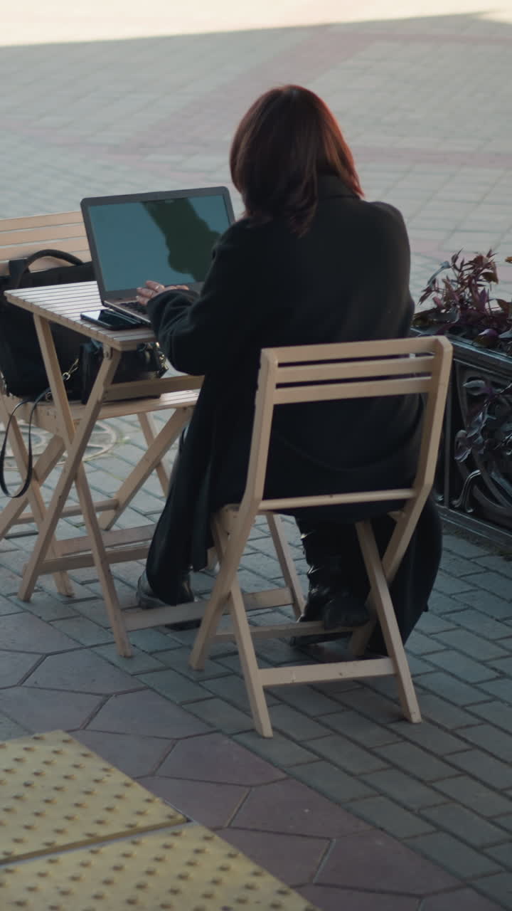 mujer profesional escribiendo en una computadora portátil al aire libre, con el reflejo del poste visible en la pantalla, trabajando en un entorno urbano en una cafetería rodeada de sillas de madera, plantas en macetas y una pasarela pavimentada